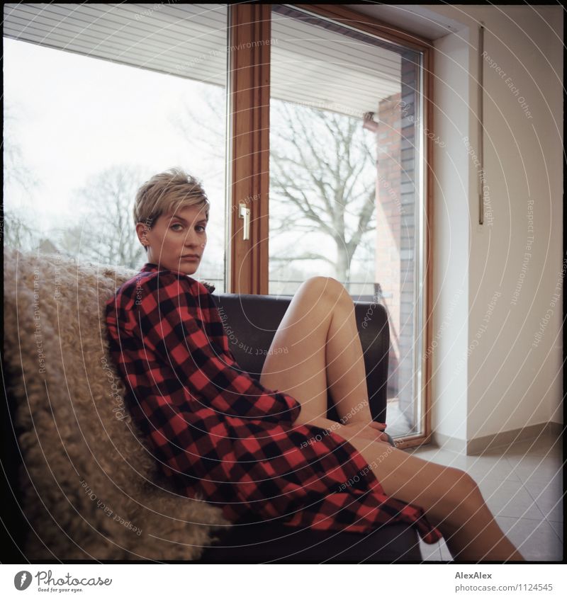 junge, sportliche Frau mit kariertem Hemd und nackten Beinen sitzt auf einem Sofa mit Schaffell Wohnung Raum Balkontür Junge Frau Jugendliche 18-30 Jahre