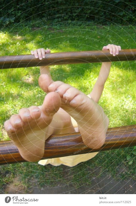 Kleines Kind hängt festgeklammert an einem Barren aus Holz, draussen in der Natur. Mädchen turnt an einer Stange, hält sich mit den Händen fest und streckt die nackten Füße nach oben. Lustige Füße, wirken durch Perspektive überdimensional groß und skurril.