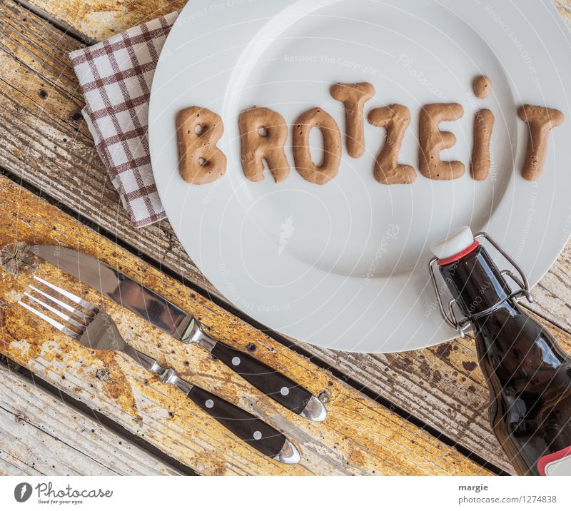 :Auf einem rustikalen Holztisch ein Teller mit den Buchstaben BROTZEIT, Messer und Gabel, Serviette und eine Flasche Bier Lebensmittel Brot Vesper Ernährung