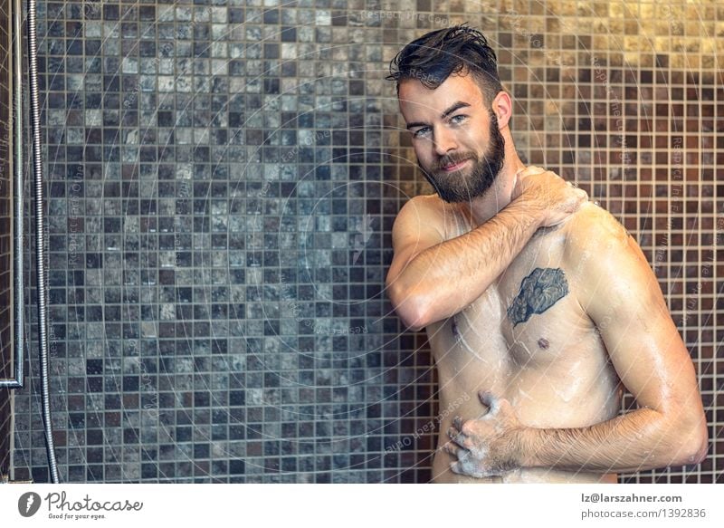 Freundlicher junger Mann, der unter der Dusche sich einseift Haut Gesicht Wellness Bad Erwachsene Vollbart Lächeln nass Sauberkeit vor der Kamera Baden Pflege