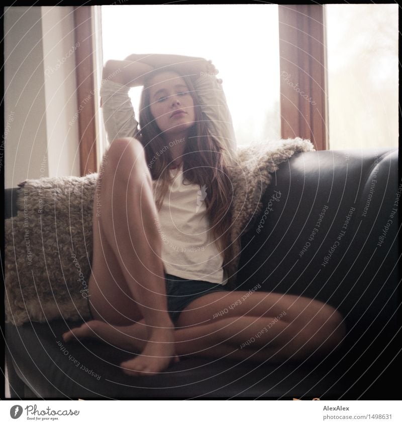 Portrait - Junge, langbeinige Frau sitzt in Shirt und Hotpants auf einem braunen Sofa mit Schaffell und schaut herausfordernd in die Kamera Lifestyle Stil