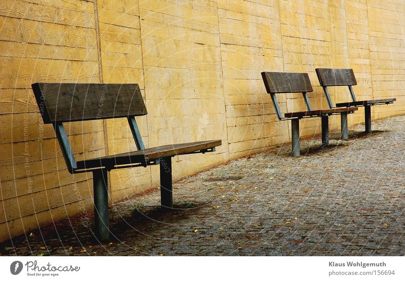 3 Sitzbänke auf gepflasterem Untergrund vor einer gelben Mauer aus Beton Bank Wand ruhig Schatten Einsamkeit nutzlos Kopfsteinpflaster Bürgersteig Park