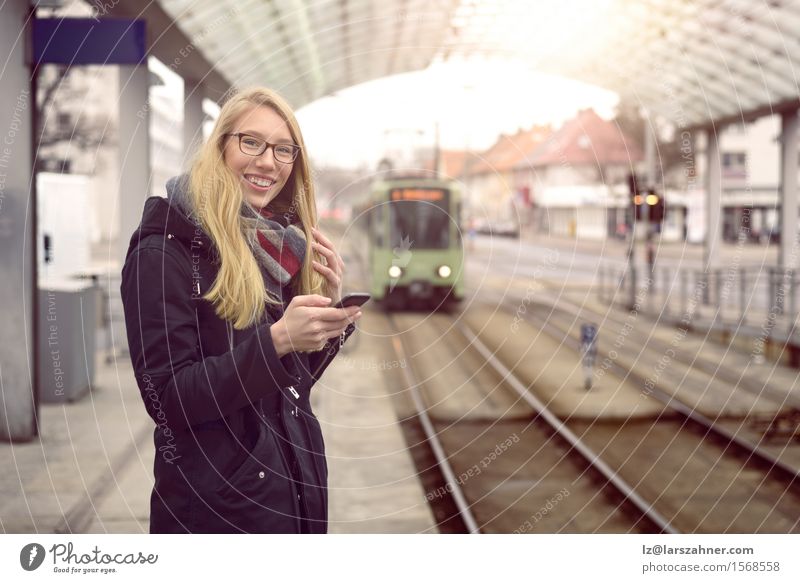 Lächelnde junge Frau in schwarzem Mantel, blonden Haaren und Brille steht am Bahnhof Glück Ferien & Urlaub & Reisen Winter Telefon Erwachsene Verkehr Eisenbahn