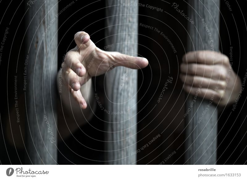 Hände eines eingesperrten Menschen klammern sich an dicke Holzstäbe im Gefängniss und sind Hilfe suchend nach draussen ausgestreckt  um Gnade flehend Hand