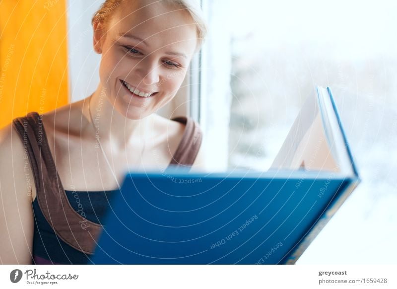 Hübsche junge lächelnde Frau liest ein Buch mit blauem Einband, während sie am Fenster sitzt Lifestyle Glück schön Erholung lesen Haus Bildung Schule lernen