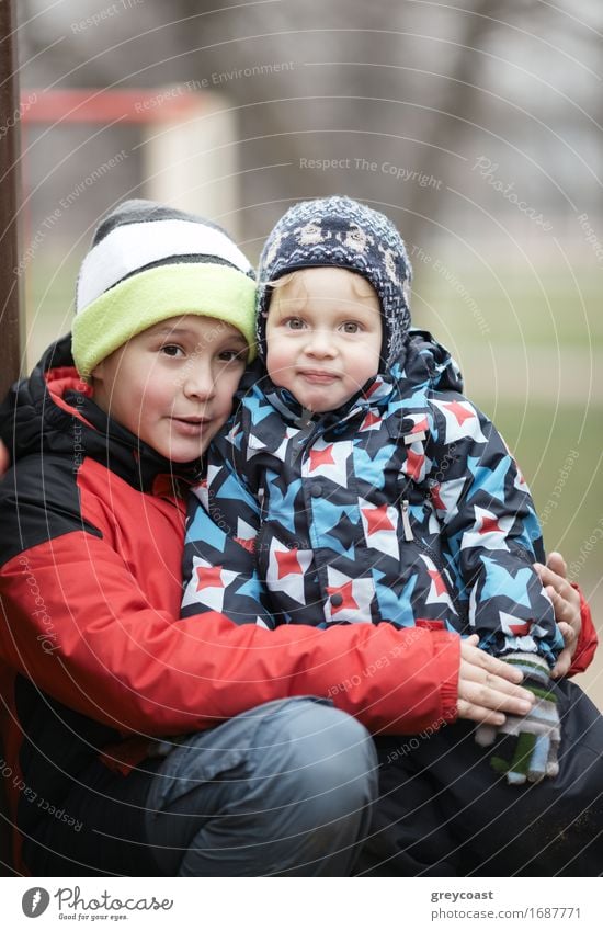Zwei entzückende junge Brüder im Freien im Winter warm eingepackt gegen das kalte Wetter mit dem älteren Jungen kuscheln sein Kleinkind Geschwister auf dem Schoß