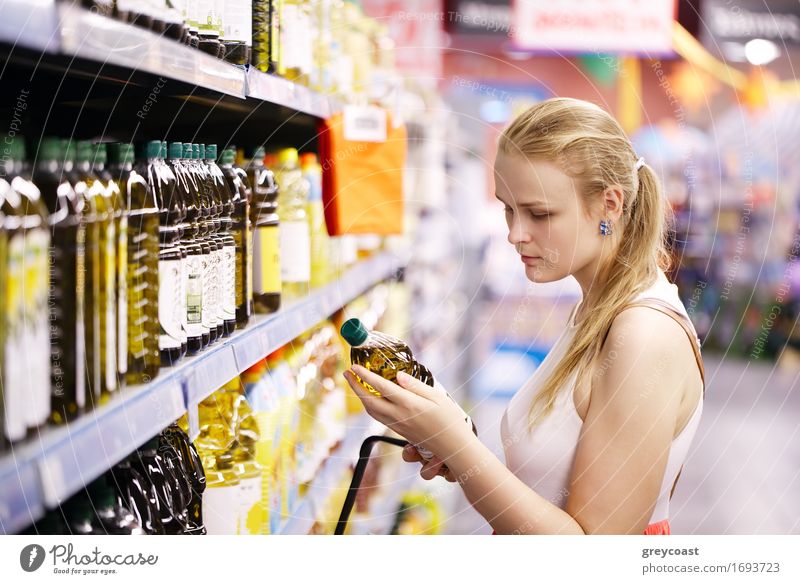 Junge blonde Frau, die eine Olivenölflasche aus den Regalen eines Supermarktes nimmt und das Etikett liest Flasche kaufen Junge Frau Jugendliche Erwachsene 1