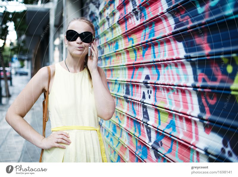Frau mit Sonnenbrille, die sich mit einem Mobiltelefon unterhält, während sie an einer mit buntem Graffiti oder einem Wandgemälde bedeckten Wand steht Lifestyle