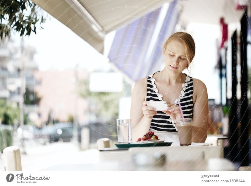 Attraktive Frau, die ein Gebäck mit ihrem Smartphone fotografiert, während sie an einem Tisch in einem Open-Air-Restaurant sitzt und Erfrischungen genießt