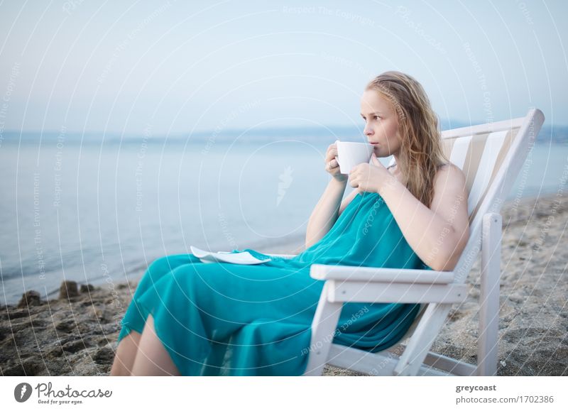 Weite Aufnahme eines jungen Mädchens, das eine Tasse Tee am Meer genießt, auf einem Liegestuhl sitzend und mit einem glückseligen Gesichtsausdruck mit Blick auf einen tropischen Strand