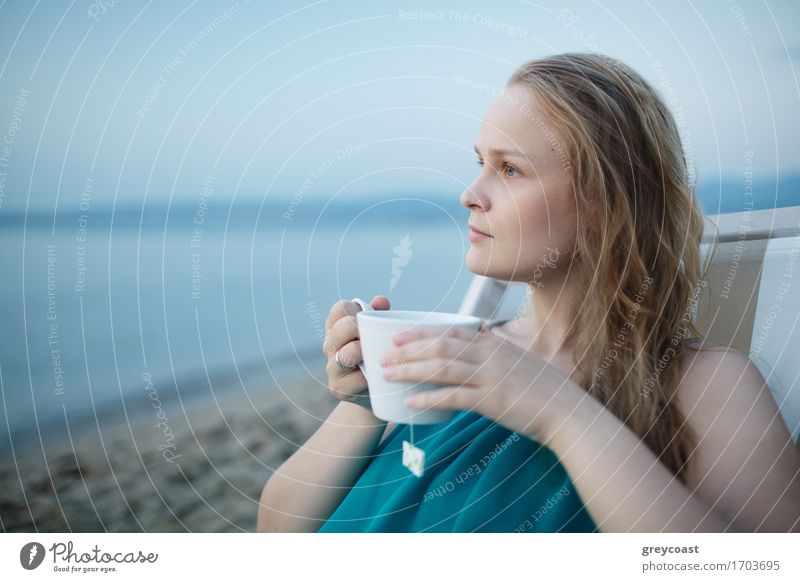 Frau, die eine Tasse Tee am Meer genießt, sitzt entspannt auf einem Liegestuhl mit einem glückseligen Ausdruck mit Blick auf einen tropischen Strand Getränk