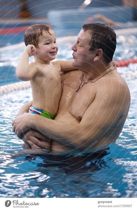 Großvater spielt mit seinem kleinen Enkel in einem Schwimmbad und hält ihn über dem Wasser in seinen Armen, während sie sich gegenseitig anlächeln und einen zärtlichen Moment teilen