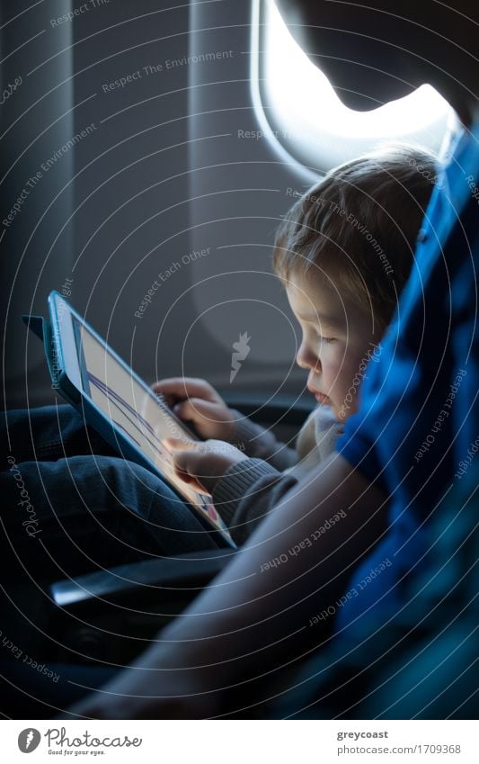 Kleiner Junge sitzt während eines Fluges in seinem Sitz und spielt zufrieden mit einem Tablet-Computer in einem Flugzeug, beobachtet von seiner Mutter