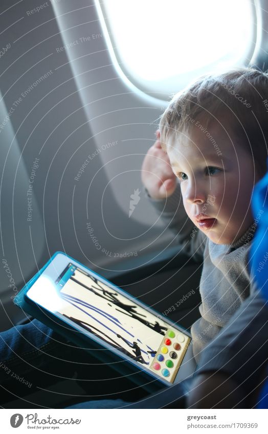 Kleiner Junge sitzt in seinem Sitz während eines Fluges und malt auf einem Tablet-Computer in einem Flugzeug Freizeit & Hobby Spielen Computerspiel