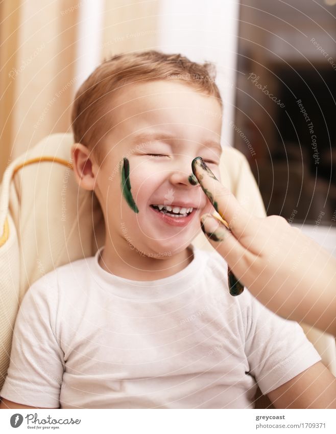 Kleiner Junge lacht, als seine Mutter malt sein Gesicht Streifung seine Wange und tupft seine Nase mit schwarzer Farbe Gesicht Freude Glück Freizeit & Hobby