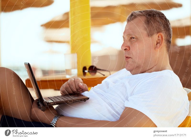 Mann, der sich mit einem Laptop in einem Strandresort auf einem bequemen Stuhl unter einem Sonnenschirm aus Stroh entspannt und nachdenklich in die Ferne starrt, Seitenansicht