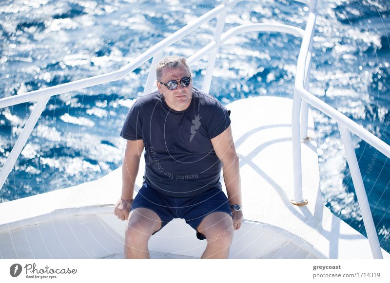 Hohe Winkel Ansicht eines Mannes mittleren Alters mit Sonnenbrille sitzt auf dem Bug eines Bootes an einem heißen Sommertag mit dem funkelnden Meer unten