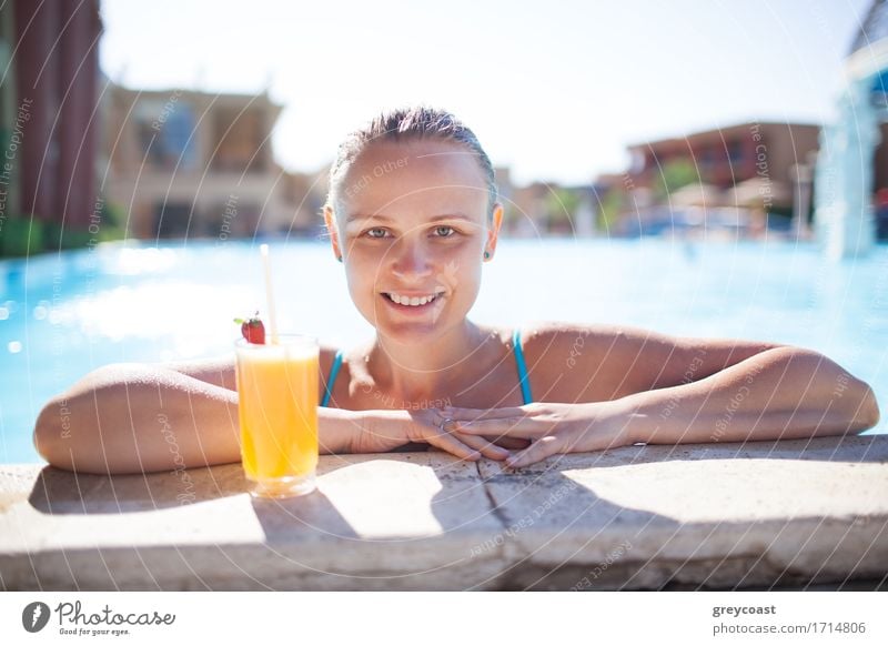 Lächelnde junge Frau, die ein erfrischendes Orangengetränk oder einen Cocktail im Pool genießt und ihre Arme auf die Poolumrandung stützt und in die Kamera lächelt