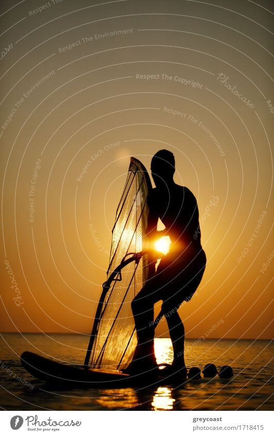 Mann sailboarding bei Sonnenuntergang Silhouette gegen die helle Kugel der Sonne in einem bunten orange Himmel auf einem ruhigen Meer Erholung