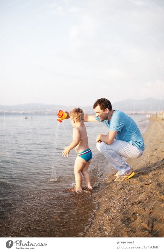 Junger Vater spielt mit seinem Sohn am Strand und kauert sich an den Rand des Wassers, während das Kleinkind fröhlich im seichten Wasser paddelt und mit einer Gießkanne spielt