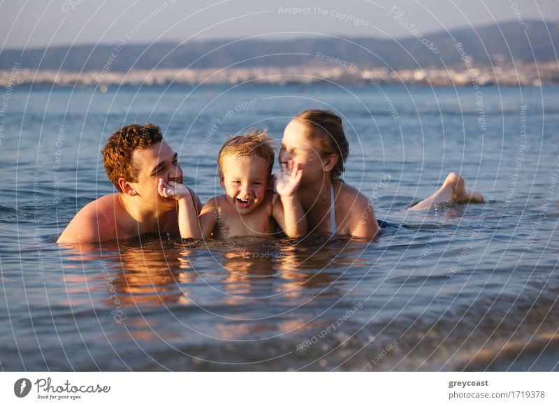 Lachender kleiner Junge flankiert von seinen liebenden Eltern, die gemeinsam im seichten Wasser am Rande des Meeres paddeln Lifestyle Freude Glück schön Leben