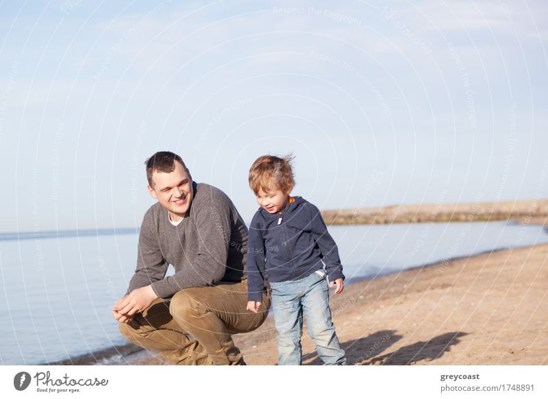 Lächelnder Vater mit seinem kleinen Sohn am Strand, der sich lächelnd in den Sand hockt, während der Junge neben ihm steht Freude Glück Meer Erwachsene