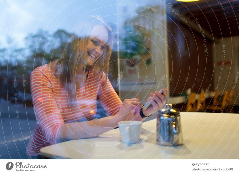 Blick durch das Glasfenster auf eine junge Frau, die in einer Cafeteria ein Mobiltelefon benutzt und lächelnd eine Textnachricht liest Heißgetränk Kaffee Tee