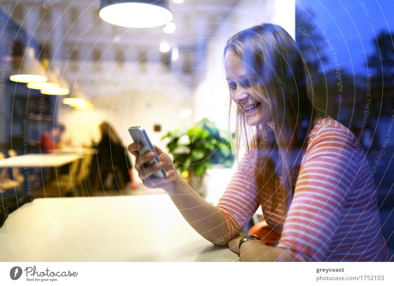 Blick durch das Glasfenster mit Reflexionen einer jungen Frau, die in einem Restaurant sitzt und mit einem Handy eine Textnachricht mit einem Lächeln liest
