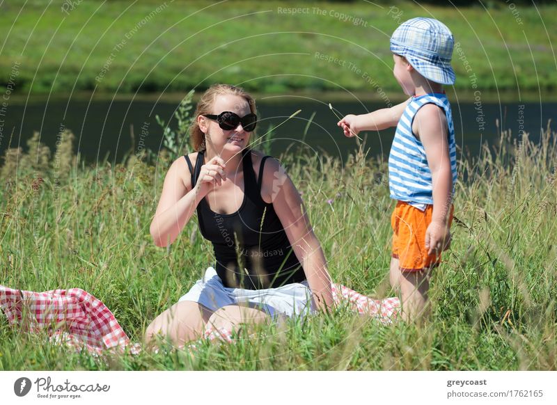 Glückliche Mutter und kleiner Sohn spielen mit Gras auf der Wiese am Ufer eines kleinen Flusses. Familie Spaß im Freien Freude Erholung Freizeit & Hobby Spielen