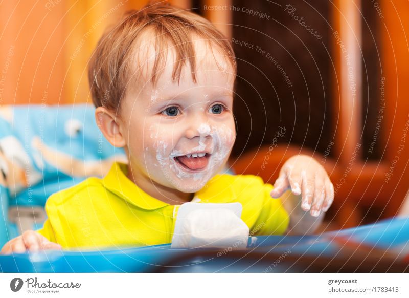 Nahaufnahme von Schuss eines glücklichen niedlichen Jungen sitzt am Futtertisch, sein Gesicht und Hände schmutzig mit Sahne Joghurt Ernährung Freude Glück Tisch