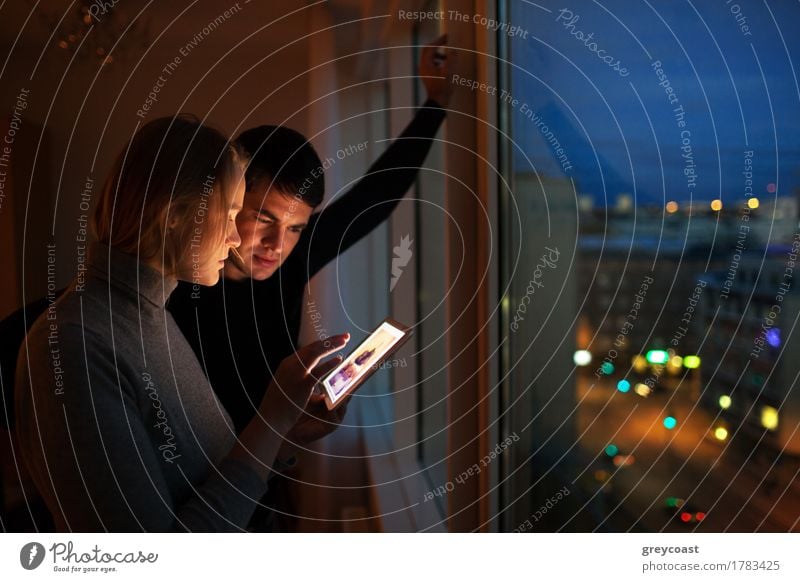 Junge Leute, die zu Hause einen Tablet-Computer benutzen. Sie schauen sich Bilder am Fenster stehend am Abend an. Stadt Lichter im Freien Freizeit & Hobby
