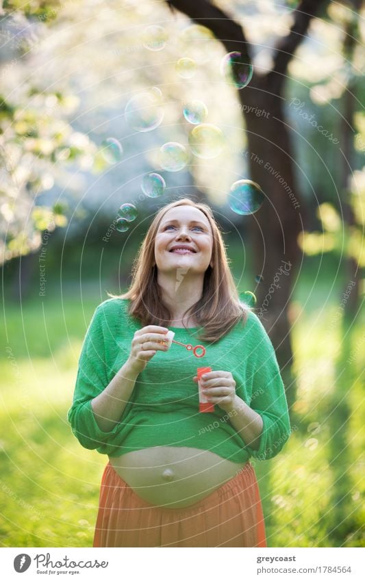 Fröhliche junge schwangere Frau genießt sorgenfreie Zeit im Park. Sie schaut auf fliegende Blasen auf dem Hintergrund der unscharfen Naturszene am sonnigen Tag