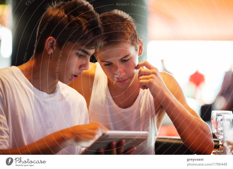 Zwei männliche Teenager surfen im Internet auf einem Tablet-Computer, während sie in einem Café sitzen. Ein junger Mann, der auf den Bildschirm zeigt