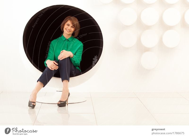 Attraktive erwachsene Frau sitzt auf kugelförmigen Stuhl mit Händen auf den Knien. Moderne weiße Wand auf dem Hintergrund Lifestyle elegant Stil Büro Mensch