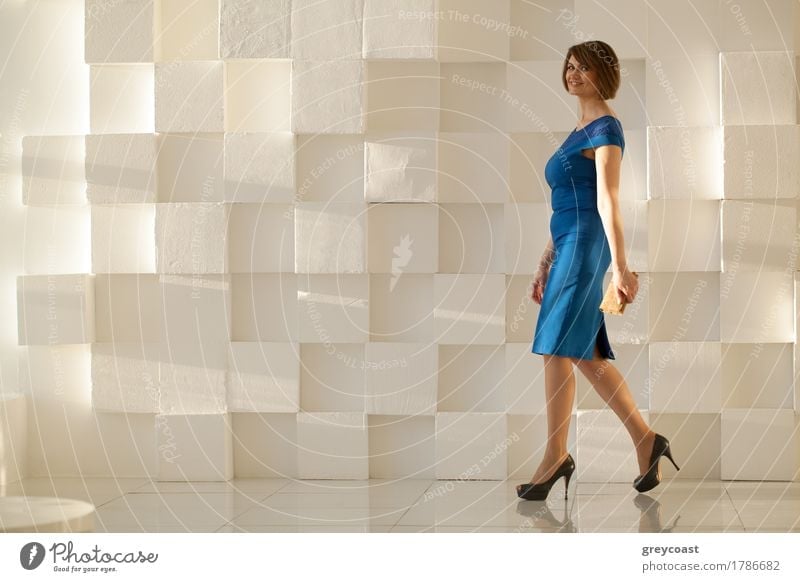 Lächelnde Frau in eleganten blauen Kleid zu Fuß gegen moderne Wand mit Brieftasche in der Hand, während der Blick auf die Kamera Lifestyle Stil Büro Mensch