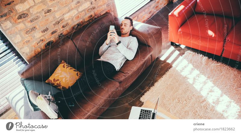 Junger erwachsener Mann, der auf das Couchgrasen mit seinem Smartphone legt Lifestyle Erholung Freizeit & Hobby Wohnzimmer Arbeitsplatz Büro Business