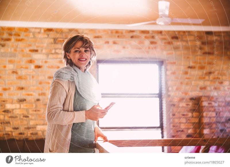 Reife Frau, die an der Kamera beim Halten von Smartphone lächelt Lifestyle Freude Häusliches Leben Wohnung Wohnzimmer Arbeitsplatz Büro Business Mittelstand