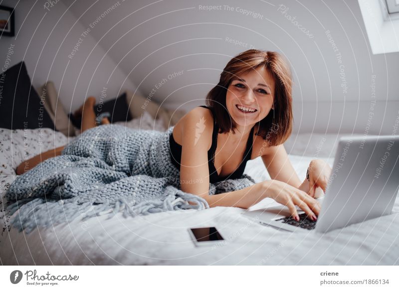 Junge kaukasische Frauen, die im Bett mit dem Laptoplächeln liegen Lifestyle Freude schön Wohnung Schlafzimmer Entertainment Schule lernen Studium Student