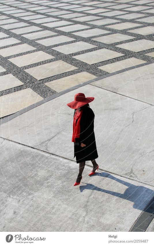 elegant gekleidete Dame mit schwarzem Mantel, rotem Hut, rotem Schal und roten Pumps spaziert auf einem großen Platz mit Beton und gemusterem Boden Mensch