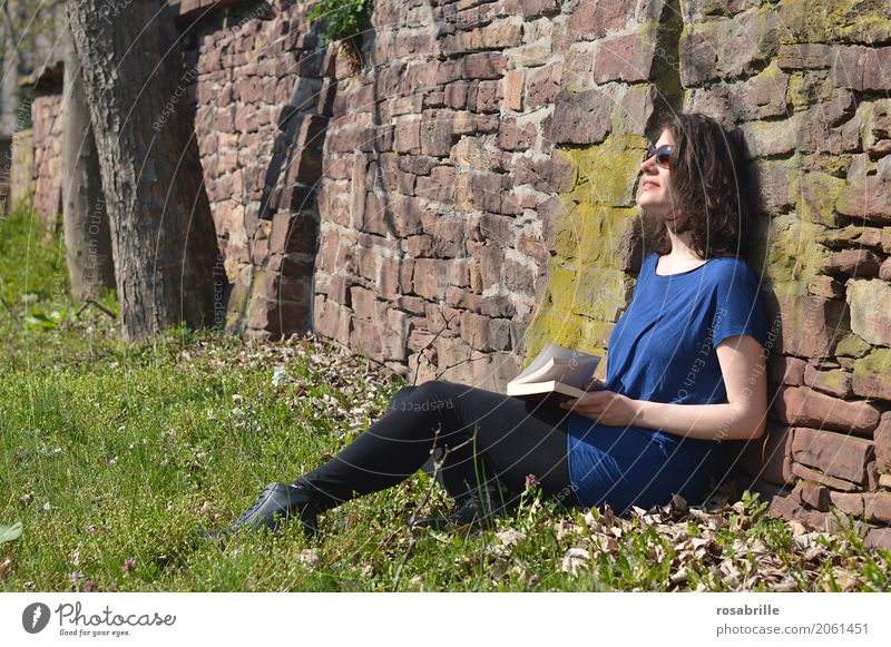 Sonne tanken - junge brünette Frau sitzt in einem Park im Gras an eine Steinmauer gelehnt mit einem Buch in der Hand und genießt die Sonne Mensch feminin