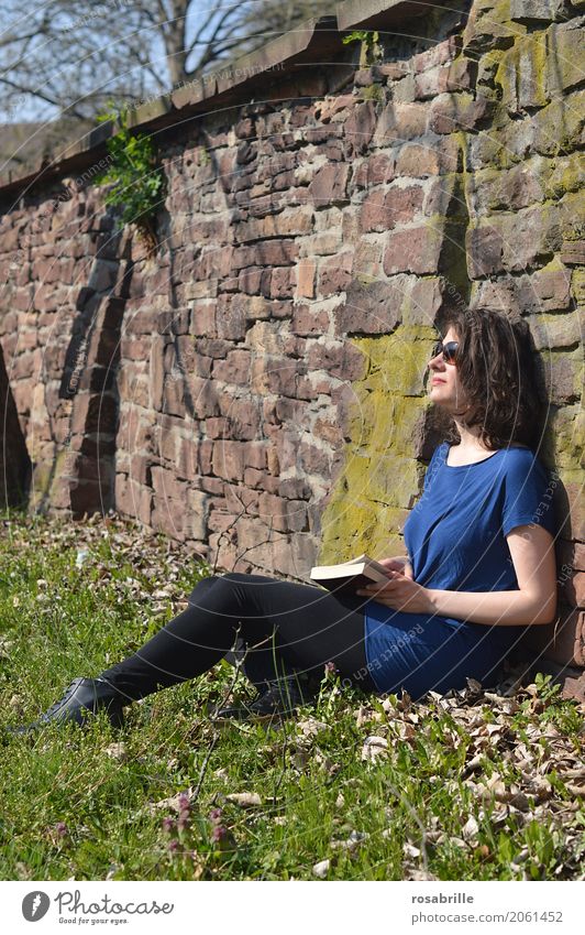 Sonne tanken - junge brünette Frau sitzt in einem Park im Gras an eine Steinmauer gelehnt mit einem Buch in der Hand und genießt die Sonne Wohlgefühl