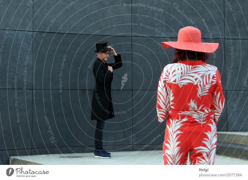 eleganter Herr in schwarzem Gehrock und Zylinder steht vor einer dunkelgrauen Wand, im Vordergrund Rückansicht einer Frau mit rot-weißem Overall und rotem Hut