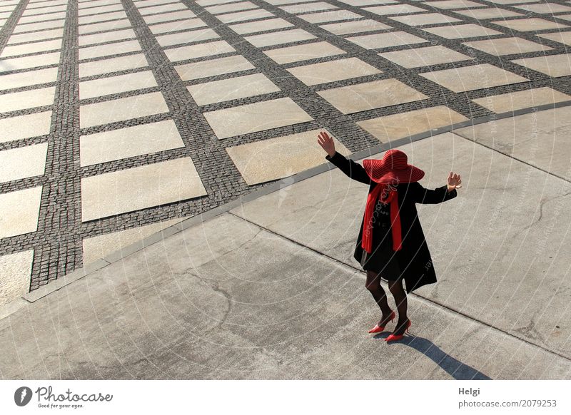 elegant gekleidete Dame mit schwarzem Mantel, rotem Hut, rotem Schal und roten Pumps tanzt auf einem großen Platz mit Beton und gemustertem Boden Mensch feminin