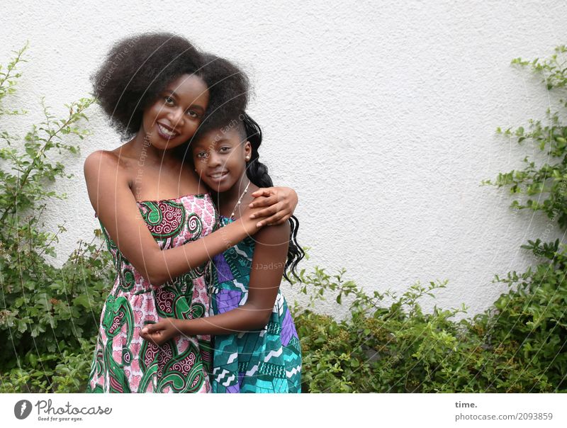 Arabella und Gloria feminin Mädchen Frau Erwachsene Schwester 2 Mensch Grünpflanze Mauer Wand Kleid Schmuck schwarzhaarig brünett langhaarig Locken Afro-Look
