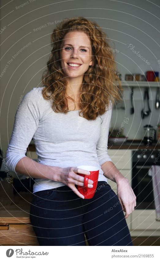 junge, rothaarige, schöne Frau mit Locken und Sommersprossen in ihrer Küche mit roter Tasse in der Hand, lächelt in die Kamera Kaffee Küchentisch