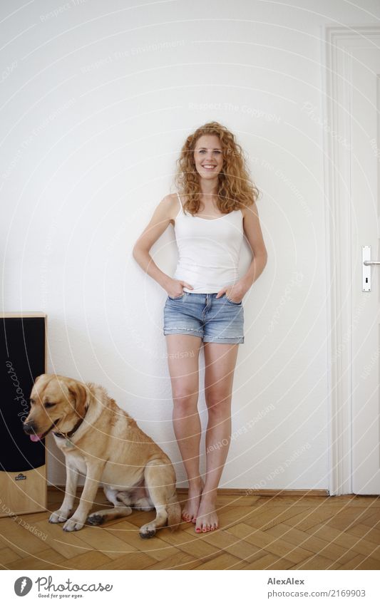 junge, rothaarige Frau mit Locken und Sommersprossen steht barfuß in Hot Pants vor einer weißen Tür und lächelt, neben ihr ein blonder Labrador Lifestyle Freude