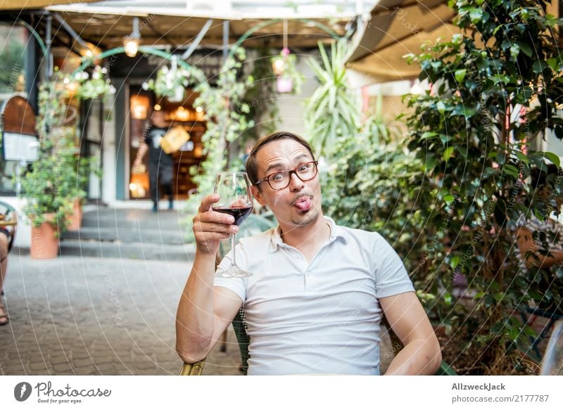 Mann mit Weinglas steckt die Zunge raus 1 Mensch Junger Mann sitzen Tag Außenaufnahme Wärme Sommer Rotwein Alkohol trinken Zuprosten Restaurant Terrasse Freude