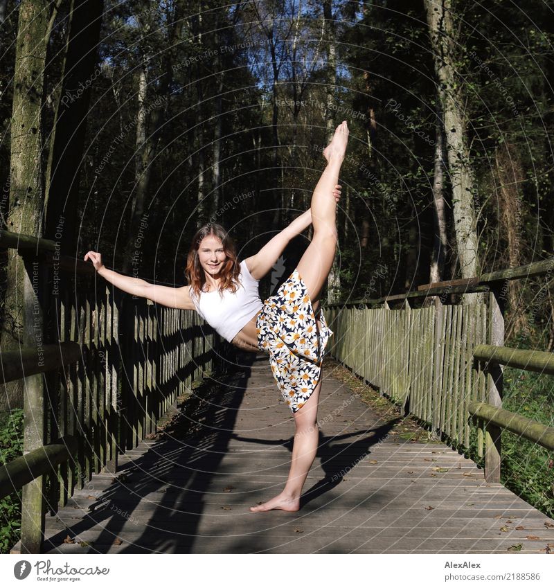 Junge, sehr sportliche und muskulöse Frau steht auf einer Holzbrücke im Wald und macht eine Tanzpose, bei der sie ein Bein über den Kopf gestreckt hoch hält, sie hat ein bauchfreies T-Shirt an und ist barfuß