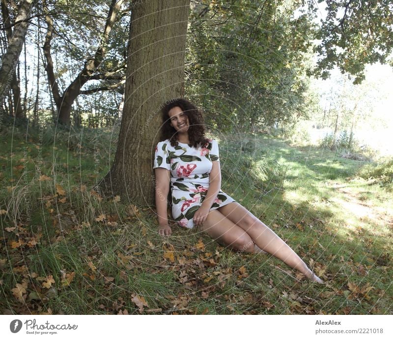 Ganzkörperbild einer großen, schönen Frau mit langen, dunklen, lockigen Haaren in der Natur die barfuß unter einem Baum sitzt Freude Leben harmonisch Ausflug