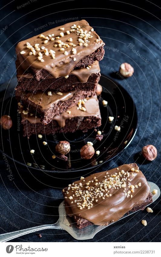 ..duftet nach Haselnuss - aufgetürmter Schokoladenkuchen mit einem Kuchenstück auf Tortenheber vor schwarzem Hintergrund Krokant Haselnusscreme Brownie